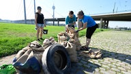 Auf dem Foto sind drei Menschen, die Müll in Säcke packen. Vor ihnen stehen bereits zahlreiche volle Säcke und auch ein Autoreifen.