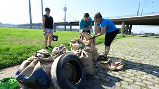 Auf dem Foto sind drei Menschen, die Müll in Säcke packen. Vor ihnen stehen bereits zahlreiche volle Säcke und auch ein Autoreifen.