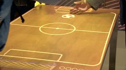 Auf dem Foto ist ein Tisch, auf dem Hände per Touchscreen ein Bild eines Balls bewegen.