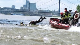 Auf dem Foto ist ein Mann, der auf dem Wasser treibt. Direkt neben ihm springt jemand von einem Boot der Feuerwehr in Tauchermontur ins Wasser.