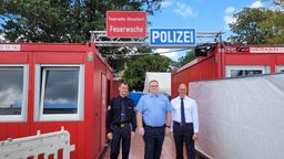 Drei Männer in verschiedenen Uniformen, über ihnen zwei Schilder mit Feuerwache und Polizei