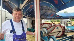 Mann mit blauer Latzhose lehnt an einem Pfosten einer Raupenbahn