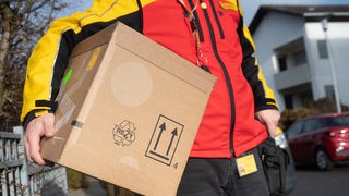 Auf dem Foto ist ein Mann in typischer rot-gelber DHL-Kleidung, der ein großes Paket unter dem Arm trägt.