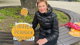 Ehrenamtlerin Hella Henckel sitzt auf einer Zuhörbank in Düsseldorf-Flingern und hält ein Schild mit dem Namen der Initiative in der Hand