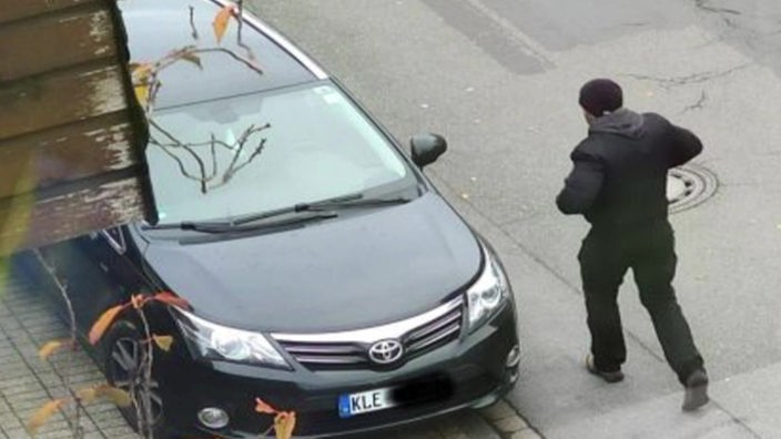 Ein Maskierter Täter steht vor einem schwarzen Auto.