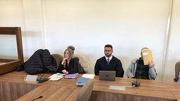 Eine Anwältin und ein Anwalt sitzen im Gerichtssaal neben einem Mann der eine Mappe vor sein Gesicht hält