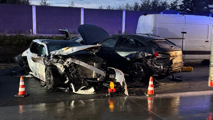 Auf dem Foto ist ein komplett zerstörter weißer Sportwagen, daneben ein schwarzer Audi mit offener Motorhaube und zerstörtem Heck.