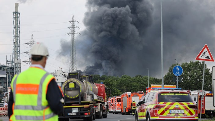 Einsatzfahrzeuge der Feuerwehr stehen unweit einer Zufahrt zum Chempark Leverkusen, über dem eine dunkle Rauchwolke aufsteigt.