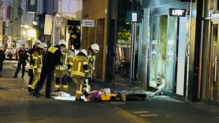 Polizisten und Feuerwehrleute untersuchen vor dem Geschäft Trümmerteile.