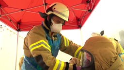 Ein Feuerwehrmann mit Schutzmaske hilft einem Kameraden beim Abziehen der Einsatzkleidung.