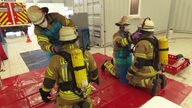 Zwei Feuerwehrleute knien vor zwei Feuerwehrkameraden. Sie bekommen Hilfe beim Ausziehen ihrer Schutzkleidung.