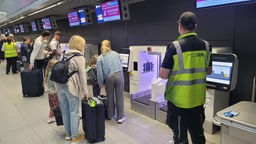 Eine Familie mit Koffern vor einem Gepäckautomaten, daneben ein Flughafenmitarbeiter in gelber Warnweste