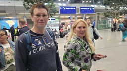 Ein Mann mit Pulli und Rucksack, daneben eine Frau mit Handy in der Hand in der Abflughalle