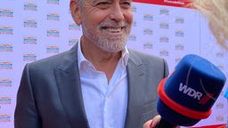 Man sieht George Clooney, wie er lächelnd vor einem Mikrofon steht. Hinter ihm ein roter Teppich. 