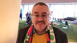 Ein Mann mit Schnäuzer, großer Brille und grauem Haarzopf trägt einen bunten Fan-Schal um den Hals