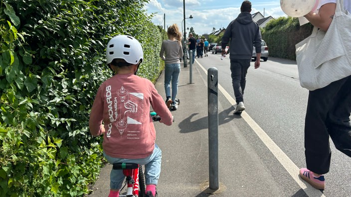 Ein Kind auf einem Fahrrad befindet sich auf einem Gehweg. Neben ihm und vor ihm laufen Personen.