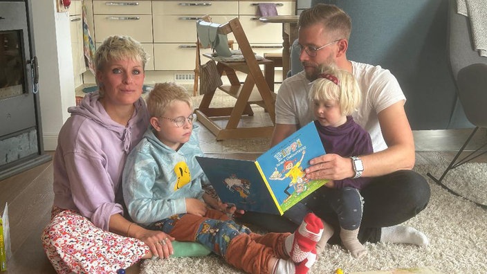 Auf dem Foto sind eine Frau und ein Mann, die jeweils ein Kind auf dem Schoß haben. Der Mann liest gemeinsam mit den Kindern ein Buch.