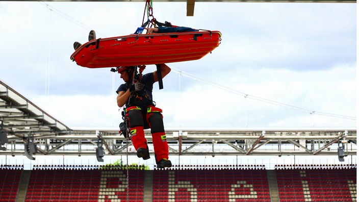 Höhenretter und Testperson in einer Liege seilen sich vom Dach des Stadions ab