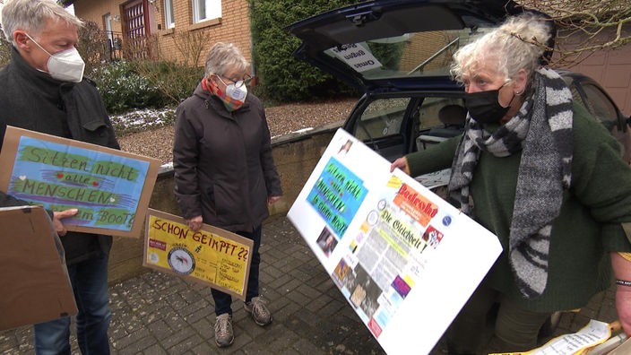 Zwei Frauen und ein Mann stehen vor einem Auto mit geöffneter Heckklappe und halten Demo-Plakate in den Händen