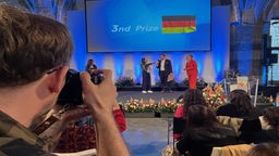 Ein Fotograf fotografiert mehrere Personen auf einer Bühne im Krönungssaal des Aachener Rathauses. Über ihnen ist eine große Tafel mit der Aufschrift "3nd Prize".