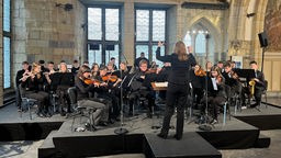 Schüler und Schülerinnen sitzen auf einer Bühne im Aachener Rathaus und spielen auf Streichinstrumenten, vor ihnen steht die Dirigentin.