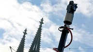 Eine Videokamera, im Hintergrund der blaue Himmel und die Spitzen des Kölner Doms