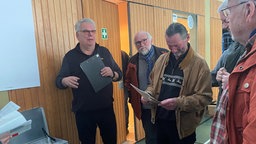 mehrere Männer im Gespräch in einer Turnhalle, halten Listen in der Hand