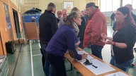 Frauen unterhalten sich in einer Turnhalle, zwischen ihnen steht eine Bierbank mit Papier und Stiften