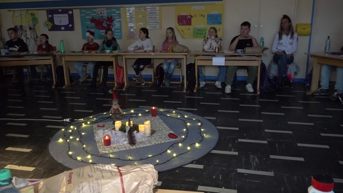 Kinder sitzen an ihren Tischen in dem Klassenraum im Kreis, in der Mitte Kerzen und weihnachtliche Dekoration.