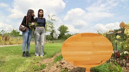Zwei Frauen stehen mit Blumen vor einem schlichten Grab mit Holztafel