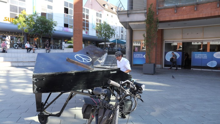 Mann mit Hut sitzt am Klavier, das auf einem Platz in der Innenstadt steht