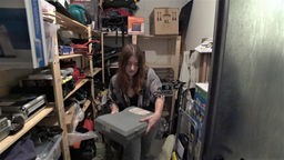 Eine junge Frau hockt in einem engen Kellerraum und hält eine Kiste.