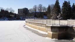 Eine vollgeschneite Talsperre, dahinter befinden sich zahlreiche Bäume so wie Wohnhäuser.