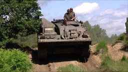 Auf dem Foto ist ein Panzer mit zwei Insassen, der über einen Weg zwischen Bäumen und Wiesen entlangfährt.
