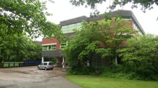 Auf dem Foto ist das Maria-Sibylla-Merian-Gymnasium: ein grau-braunes Betongebäude mit grünen Fensterrahmen.