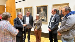 Auf dem Foto sind mehrere Personen, die den NRW-Innenminister Herbert Reul anschauen. Dieser hält eine Mappe in der Hand.