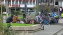 Auf dem Foto sind Männer mit dicken Jacken, die auf den Sitzgelegenheiten am Krefelder Theaterplatz sitzen, daneben sind viele Säcke und ein Fahrrad mit Decken darauf.
