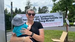 Alice König steht mit ihrem Sohn Anton auf dem Arm vor der GFO-Klinik Bad Honnef. hinter ihr ein Plakat, dass mehr Geburtsstationen in der Region fordert.