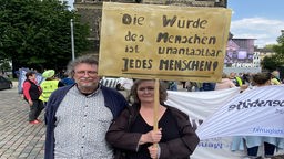 Eine Frau und ein Mann stehen vor einer Kirche. Die Frau hält ein Schild mit der Aufschrift "Die Würde des Menschen ist unantastbar. Jedes Menschen." hoch.