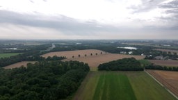 Auf dem Foto ist eine Aufnahme einer Landschaft mit Feldern, Wald und einem See aus der Vogelperspektive.