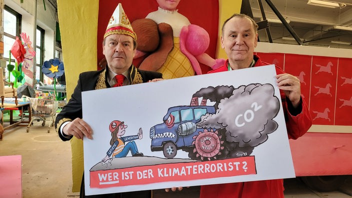 Zwei Männer, einer in Karnevalsuniform, einer im roten Overall, halten zusammen ein Plakat hoch