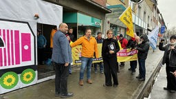 Mona Neubaur und Omid Nouripour stehen auf einem Gehweg, im Hintergrund hält eine kleine Gruppe eine gelbe Flagge