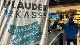 In einem Supermarkt in Kempen hat eine neue "Plauderkasse" eröffnet. Zu sehen ist die Kasse mit einer Beach-Flag davor, auf der "Plauderkasse, hier haben sie alle Zeit der Welt" steht.