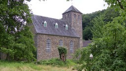 Ein altes Kirchengebäude aus braunen Steinen und einem kleinen Kirchturm ist von Bäumen umrundet.