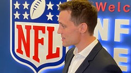 Andreas Steinforth steht vor einem leuchtenden NFL Wappen. Auf dem Wappen sind ein weißer Football und sechs weiße Sterne zu sehen.