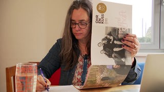 Ein Frau mit langem, dunklem Haar und Brille hält in einer Hand hochkant ein Print-Magazin mit dem Titel "Ohrenkuss". Mit der anderen Hand schreibt sie etwas auf ein weißes Blatt Papier. 