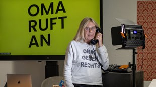 Eine Oma steht vor einem grünen Hintergrund mit "Oma ruft an"-Aufschritt und hält einen Telefonhörer in der Hand