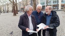 Drei grauhaarige Männer reden miteinander und halten Broschüren in den Händen