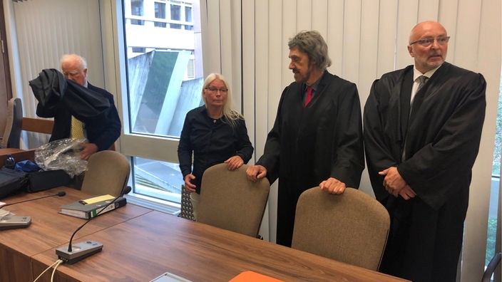 Frau mit weißen Haaren im Gerichtssaal neben Anwälten in schwarzer Robe
