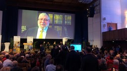 Ein Saal voller Menschen, Menschen  auf einer Bühne stehend und das Bild von Pinchas Goldschmidt auf einer Leinwand projeziert im Hintergrund